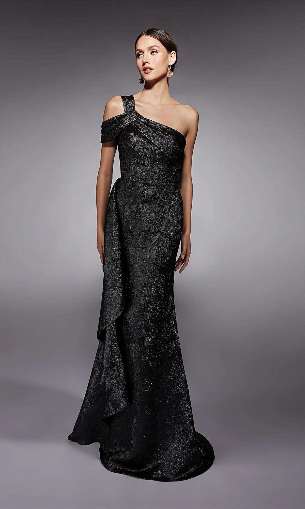 A woman wears the Alyce 27734 Long Formal Dress in Dripping Diamonds (Silver), a floor-length, one-shoulder gown with draped detail, ideal for a mother of the bride, standing against a plain gray background.